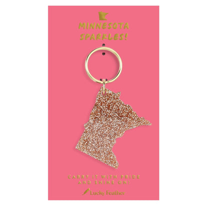 A glittery Minnesota state-shaped keychain with a gold-tone ring, displayed on a pink backing card with gold text that reads 'MINNESOTA SPARKLES!' and 'Lucky Feather'.