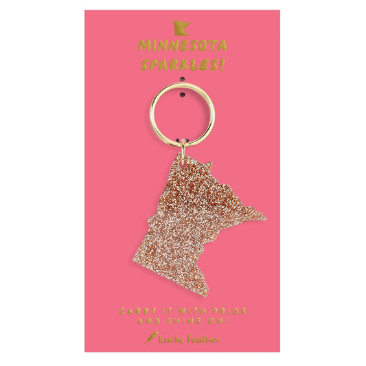 A glittery Minnesota state-shaped keychain with a gold-tone ring, displayed on a pink backing card with gold text that reads 'MINNESOTA SPARKLES!' and 'Lucky Feather'.