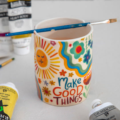 A multicolored ceramic painter's cup with a notch for holding a paintbrush, displayed alongside various paintbrushes and art supplies.
